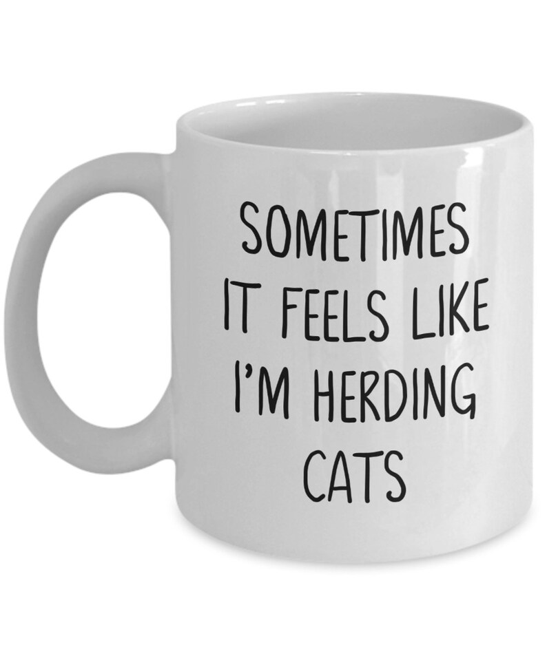 Może przedstawiać: Biały ceramiczny kubek do kawy z czarnym tekstem, kt&oacute;ry brzmi "Sometimes it feels like I'm herding cats".