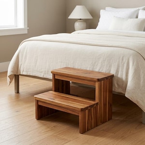 May include: A two-step wooden stool featuring a natural brown wood grain finish with rounded edges on each step. The stool has a rectangular design with vertical side supports and a lower step positioned in front of a higher, wider top step.