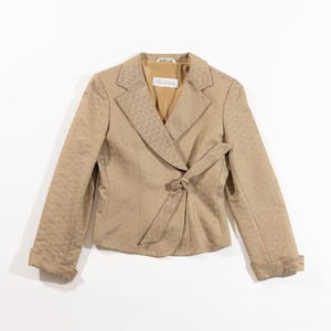 Puede incluir: Una blazer color beige con un patrón texturizado, con solapa de muesca y cierre de lazo en la cintura. Las mangas están dobladas y la blazer tiene la etiqueta "MaxMara" en el interior.
