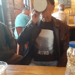 May include: A man wearing a black leather jacket and a black t-shirt with the word "NITRO" printed on it, is drinking from a white mug. He is sitting at a wooden table with a can of soda, a bag of peanuts, and a glass of peanuts on the table.