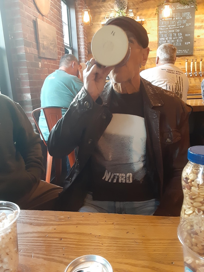 May include: A man wearing a black leather jacket and a black t-shirt with the word "NITRO" printed on it, is drinking from a white mug. He is sitting at a wooden table with a can of soda, a bag of peanuts, and a glass of peanuts on the table.