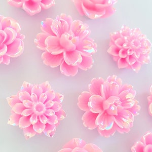 May include: Assortment of ten pink iridescent flower embellishments. The flowers are in various shapes and sizes, including roses, daisies, and carnations.