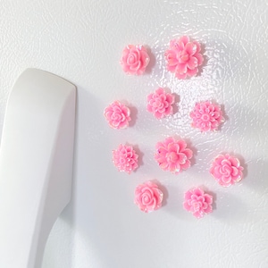 May include: A collection of ten pink flower-shaped magnets with iridescent glitter accents. The magnets are arranged on a white refrigerator.