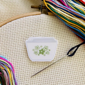 May include: A white ceramic bowl with a green floral design, a needle and thread, and a piece of Aida cloth.
