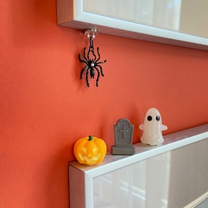 May include: Halloween-themed decorations including a black spider, an orange pumpkin, a gray tombstone, and a white ghost. The spider hangs from a clear magnet. The pumpkin has a carved face. The ghost has two black eyes.