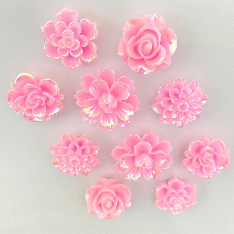 May include: Assortment of 10 pink iridescent resin flower embellishments. The flowers are in various styles, including roses, daisies, and carnations.