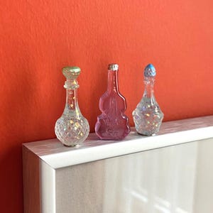May include: Three decorative glass bottles with glitter inside. The bottles have different shapes and colors, including clear, pink, and blue.