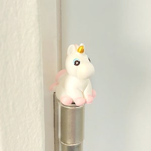 May include: A white unicorn figurine with a golden horn, pink hooves, and a pink tail sits atop a silver hinge. The unicorn has blue eyes and is positioned against a white background.