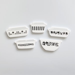 May include: Five white ceramic baking dishes with black floral and geometric designs. The dishes are arranged in a row on a white background.