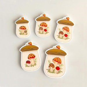 May include: Five white ceramic jars with brown lids and mushroom illustrations. The mushrooms are red, orange, and brown with white spots.