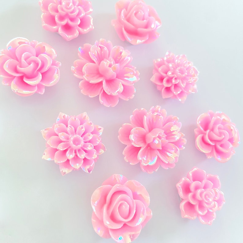 May include: Assortment of ten pink iridescent flower embellishments. The flowers are in various shapes and sizes, including roses, daisies, and carnations.