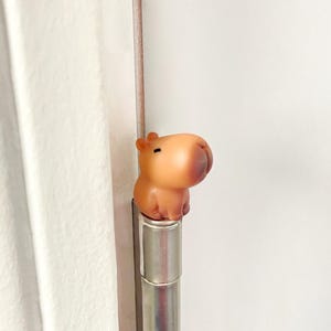 May include: A small, light brown capybara figurine with a darker brown nose and ears, perched on a silver metal rod. The figurine has a smooth, rounded shape and is set against a white background.