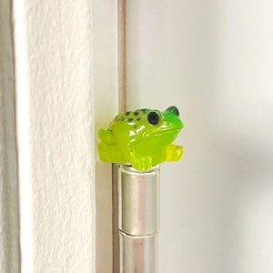May include: A green and black frog figurine with black eyes sits on top of a silver pen.