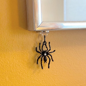 May include: A black spider charm with a white pearl in the center, hanging from a silver frame.