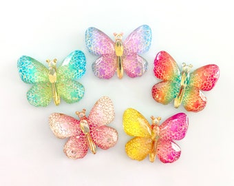 May include: Five colorful butterfly-shaped embellishments with gold accents. The butterflies are in shades of pink, blue, yellow, and green.