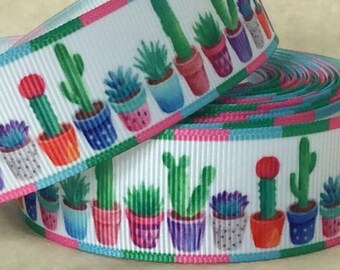 May include: A white ribbon with a repeating pattern of potted cacti in various colors and styles. The ribbon has a green, pink, and blue striped border.