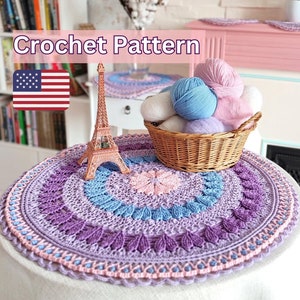 May include: Crochet pattern for a purple, blue, and pink round doily with a flower in the center. The doily is on a white table with a small Eiffel Tower figurine and a basket of yarn.