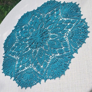 Puede incluir: Un tapete de ganchillo redondo de color verde azulado que presenta un intrincado patrón simétrico de flores y hojas con un diseño de puntadas en relieve. La pieza tiene un borde festoneado y puntiagudo, elaborado con hilo fino y una estructura calada.