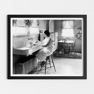 May include: A black and white photo of a woman in a kitchen, standing at the sink and washing dishes. She is wearing a white dress and has her hair pulled back. The kitchen has a tiled floor and a window with curtains.