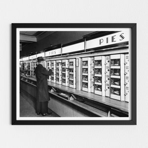 May include: Black and white framed photograph of a vintage pie vending machine. A man in a hat and coat stands in front of the machine, which is labeled "PIES" above the compartments. The photograph captures a moment in time.