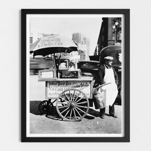 Könnte beinhalten: Schwarzweißfoto eines Straßenverkäufers, der Hotdogs, eiskalte Limonade und heiße Würstchen von einem Wagen mit einem großen Schirm verkauft, auf dem "Frankfurters and Ice Cold Lemonade" steht.