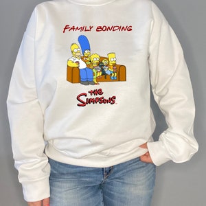 May include: White sweatshirt with a graphic of the Simpsons family sitting on a couch. The text "Family Bonding" is above the image and "The Simpsons" is below the image.