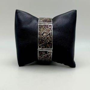 May include: A silver-toned bracelet with a rectangular, speckled, brown and gray center panel. The center panel is framed by silver filigree leaf designs. The bracelet is displayed on a black leatherette pillow.