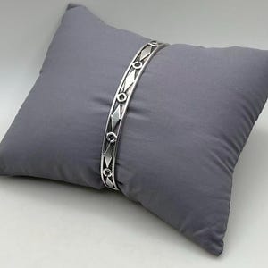 May include: A silver-colored bangle bracelet with a geometric design of diamonds and circles. The bracelet is displayed on a gray pillow, set against a white background. The bracelet's design is repeated along its length.