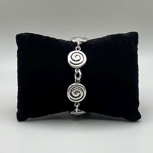 May include: A silver-toned metal bracelet featuring a series of circular spiral-shaped links connected by small jump rings. The bracelet is displayed vertically, showcasing the repeating swirl pattern of each individual metallic link component.