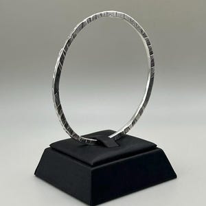 May include: A silver-colored bangle bracelet with a textured, geometric pattern. The bracelet is displayed on a black, angled display stand against a neutral background. The bangle's design features a series of small, angled cuts.