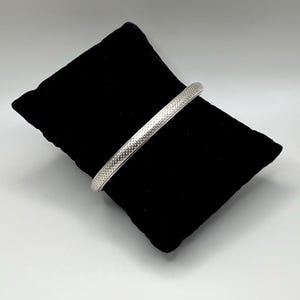 May include: A silver-colored bangle bracelet with a textured surface. The bracelet is displayed on a black velvet cushion against a white background. The bracelet's design features a repeating pattern of small, raised dots.