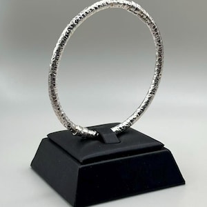 May include: A silver-colored bangle bracelet with a textured surface. The bracelet is displayed on a black square stand against a white background. The bangle is a circular shape.