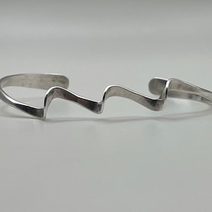 May include: A silver-toned cuff bracelet featuring a continuous, undulating wave pattern that creates a rhythmic, zigzag silhouette. The metal has a smooth, polished finish with rounded ends, forming an open-ended circular band design.