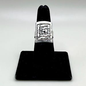 May include: A silver-toned ring with a square face featuring a hammered texture and a recessed spiral design. The ring is displayed on a black jewelry stand, showcasing its geometric pattern and metallic sheen.