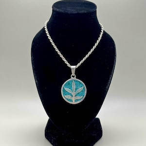 May include: Silver-toned necklace with a round pendant featuring a turquoise inlay and a leaf design. The pendant is suspended from a silver rope chain. The necklace is displayed on a black velvet bust.