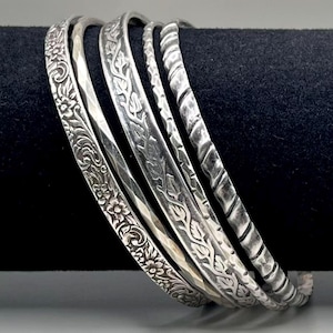 May include: A collection of silver bangles with various embossed designs. The bangles feature floral, leaf, and textured patterns. The bracelets are displayed on a black surface.