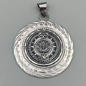May include: A circular silver pendant featuring a detailed central medallion with a sun-like face and intricate geometric patterns. The medallion is surrounded by a textured, twisted silver border and a beaded rim, topped with a triangular bail.