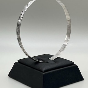 May include: A thin, circular silver bangle bracelet featuring a continuous embossed floral pattern along its outer surface. The metal has a polished, reflective finish, and the design consists of repeating flower motifs linked by delicate vine-like details.