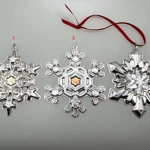 May include: Three silver snowflake ornaments with intricate designs. One has a gold center, another a clear center, and the third has a gold center. A red ribbon is attached to one.