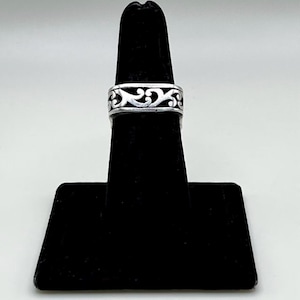 May include: A silver ring featuring a continuous band with an openwork cutout design of swirling, interconnected scroll patterns. The metal has a polished finish with darkened recessed areas that highlight the intricate, repeating filigree motif.