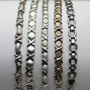 May include: Five silver-toned metal link bracelets arranged vertically and numbered one through five. The bracelets feature various designs including X-shaped links, circular discs, and interlocking geometric patterns with polished and matte finishes.