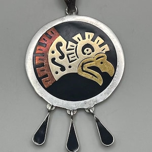 May include: A silver pendant with a circular design featuring a stylized eagle head in gold and silver against a black background. The pendant has a red and copper border and three black teardrop dangles.