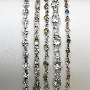 May include: Five distinct bracelets arranged vertically and numbered one through five. The first is a silver-tone chain with clear stones. The second is a gold-tone chain with heart-shaped links and multi-colored stones. The third features green oval stones. The fourth has large clear faceted stones. The fifth is a gold-tone chain with small heart charms and colorful beads.