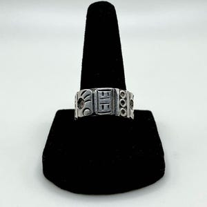 May include: A silver band ring featuring a series of embossed geometric and abstract symbols around the exterior. The metal has a polished finish with darkened recessed areas that highlight the intricate patterns and shapes engraved on the surface.