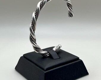 May include: A silver-colored twisted rope design cuff bracelet. The bracelet is displayed on a black jewelry stand against a white background. The bracelet's design features a braided rope pattern.