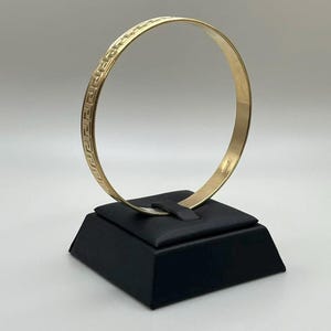 May include: A circular gold bangle bracelet featuring a continuous engraved Greek key pattern along its outer surface. The metal has a polished finish, and the band maintains a consistent width throughout its circumference, creating a classic design.
