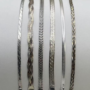 May include: Six silver-toned metal bracelets arranged vertically, numbered one through six. The collection includes various chain styles: a flat patterned link, a multi-strand braided design, a dense woven chain, a textured flat link, a smooth herringbone, and a thin braided chain.