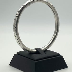 May include: A circular silver-toned bangle bracelet featuring a series of evenly spaced, etched diagonal lines along its outer surface. The bangle is displayed upright on a black square stand, showcasing its smooth, metallic finish and consistent form.