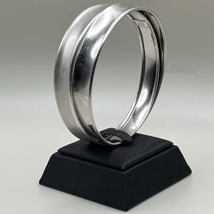 May include: A silver-toned bangle bracelet with a curved design, displayed on a black square stand. The bracelet has a smooth, polished finish, reflecting the light. The stand provides a contrasting dark base.