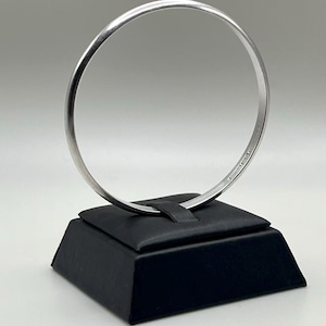 May include: A smooth, polished silver-toned bangle bracelet with a circular shape. The bracelet is displayed upright, resting in a small slot on a black, square-based stand. The metal surface reflects light, showing a clean, simple, and continuous design.